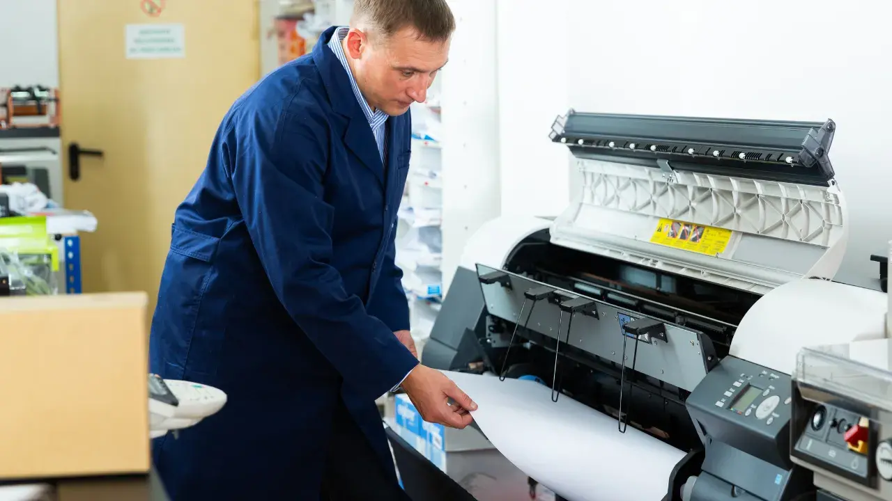 Una mano tirando cuidadosamente de un papel atascado en un plotter, mostrando claramente el problema técnico.